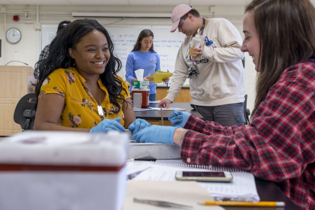 Two students sitting and talking in the Pre-College Summer Program with other students participating behind them. 